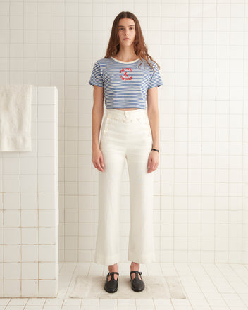 A person with long hair stands in a tiled room, wearing a striped Frog and Pony Tee - Blue and high-waisted, wide-leg pants made from a cotton rayon blend.