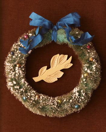 A vibrant Frond Brooch decorated with colorful ornaments and a blue ribbon, featuring a butterscotch yellow leaf accent at the center, reminiscent of vintage 1930s jewelry.