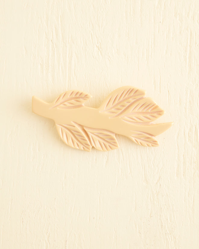 A Frond Brooch in a subtle butterscotch shade adorns the textured cream wall, reminiscent of the elegance found in 1930s jewelry.