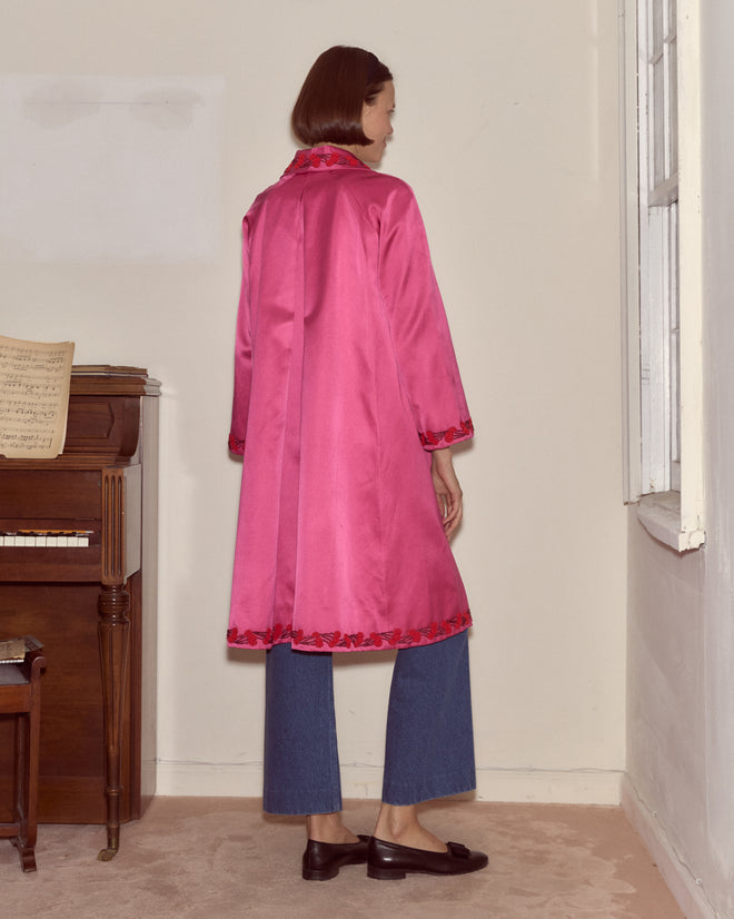 A woman wearing the Fuchsia Cherry Coat, featuring electric pink silk and hand-beaded cherry trim, pairs it with jeans as she stands by a piano and window with her back to the camera.