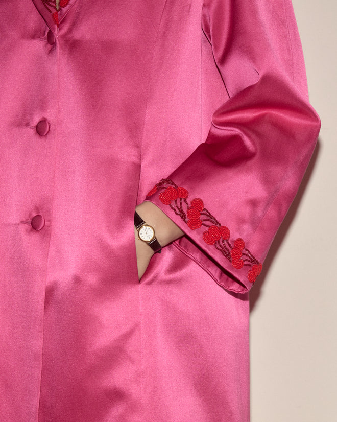 A person wearing the Fuchsia Cherry Coat—an electric pink silk coat with red embroidery and hand-beaded cherry trim—has their hand in a pocket, displaying a watch.