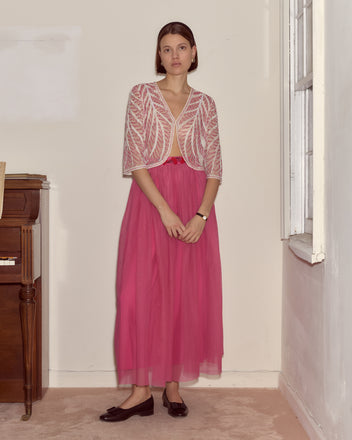 Woman in a sheer white jacket and the Fuchsia Cherry Skirt stands by a piano indoors, looking at the camera—a touch of vintage costume elegance completes the scene.