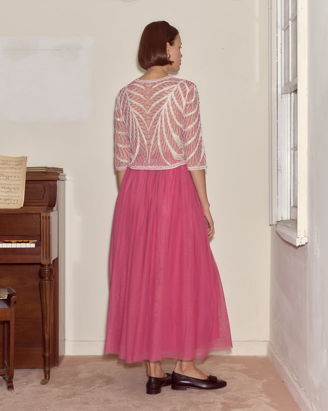 Woman wears the Fuchsia Cherry Skirt and a sheer white top, standing by a piano in a softly lit room, facing away.