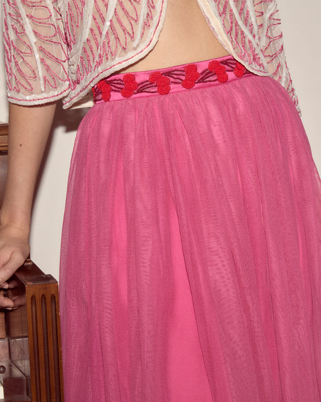 Someone wearing the Fuchsia Cherry Skirt with a red waistband and beaded cherry trim, paired with a white embroidered top, stands next to a wooden chair.
