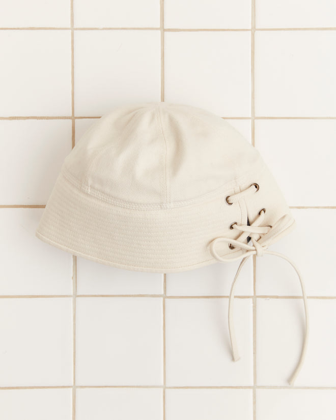 The Sailor Cap - White, crafted from cotton twill and featuring lacing detail on the side, is showcased against a white tiled background.