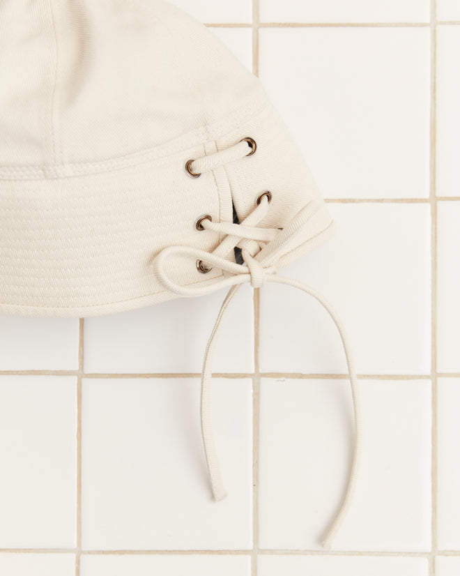 The Sailor Cap - White, crafted from cotton twill and featuring lacing detail on the side, is showcased against a white tiled background.