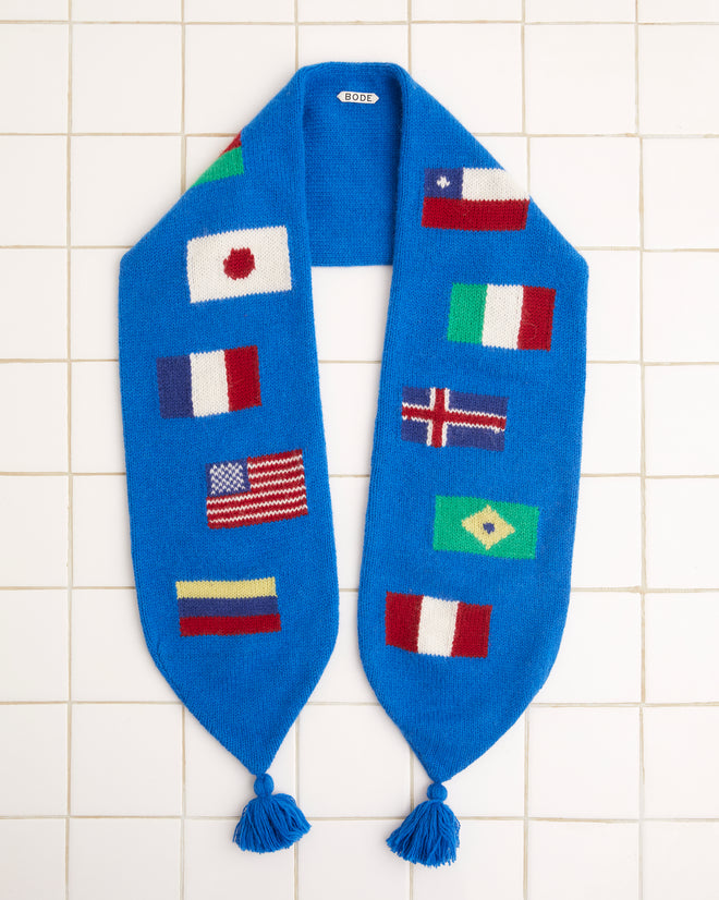 The Round-the-World Scarf - Blue, crafted from soft merino wool and featuring an intarsia flag pattern of various national flags, is elegantly displayed on a tiled surface.