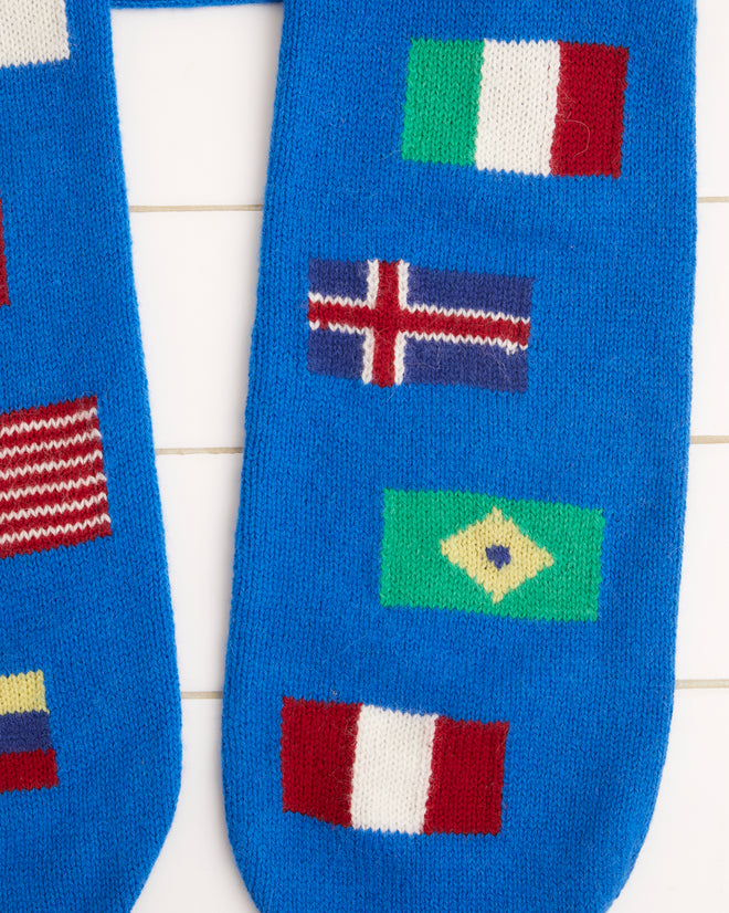 The Round-the-World Scarf - Blue, crafted from soft merino wool and featuring an intarsia flag pattern of various national flags, is elegantly displayed on a tiled surface.