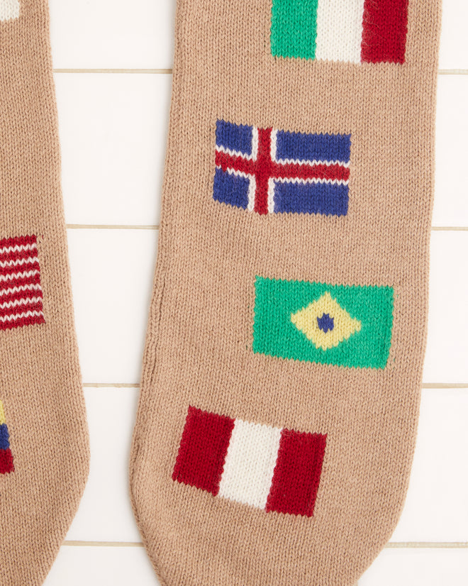 Round-the-World Scarf - Tan with an intarsia flag pattern displaying international flags such as Japan, Italy, France, USA, and Brazil on a tiled background. Made from soft merino wool for warmth and style.