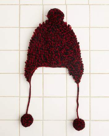 The Loopy Tassel Hat, a cozy crocheted accessory in red and black featuring ear flaps and pom-pom tassels, is artfully displayed on a white tiled background.