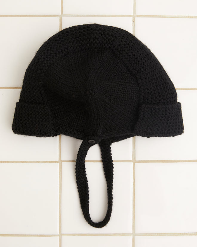 The Terrain Hat - Black, a Merino wool knit cap featuring a chin strap, is elegantly displayed on a white tiled surface, making it the perfect addition to your collection of stylish accessories.