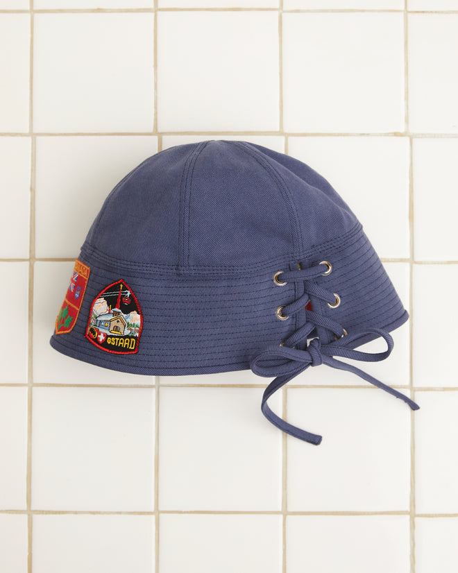 The Patched Sailor Cap - Blue, crafted from cotton twill and embellished with decorative patches and lace-up detailing, is displayed against a tiled background.