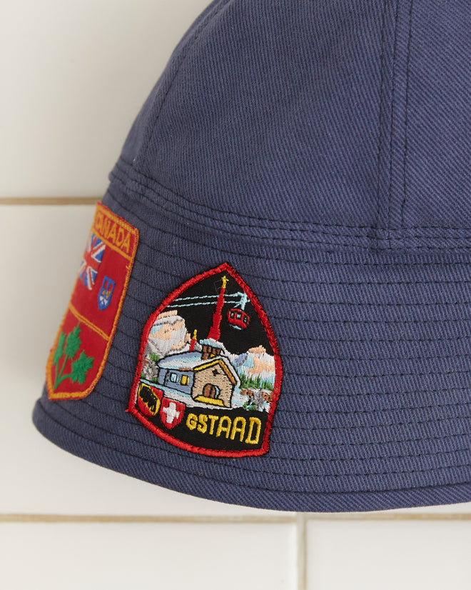 The Patched Sailor Cap - Blue, crafted from cotton twill and embellished with decorative patches and lace-up detailing, is displayed against a tiled background.