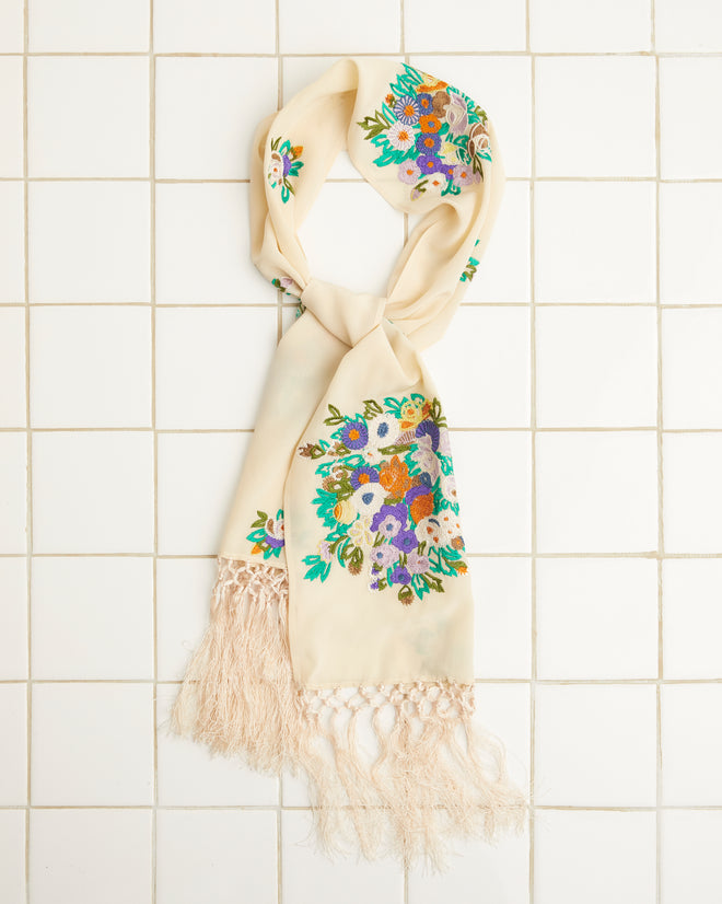 The Flower Language Scarf, made from 100% silk, showcases vibrant floral embroidery and delicate fringe tassels, beautifully displayed on a white tiled surface.