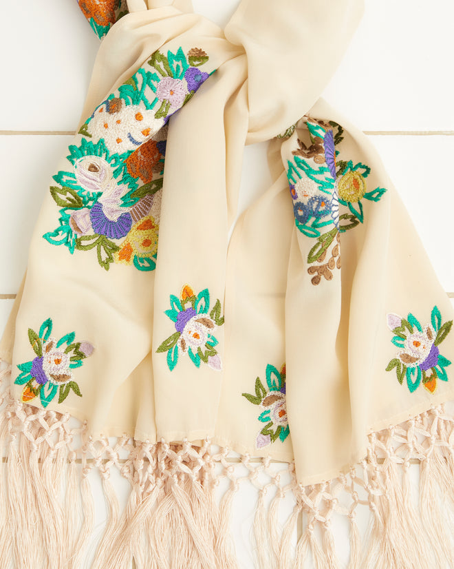 The Flower Language Scarf, made from 100% silk, showcases vibrant floral embroidery and delicate fringe tassels, beautifully displayed on a white tiled surface.