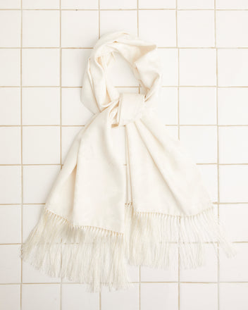 The Football Jacquard Scarf in cream, featuring a subtle football pattern and fringed edges, is elegantly displayed on a white tiled background. It is crafted from luxurious deadstock fabric.