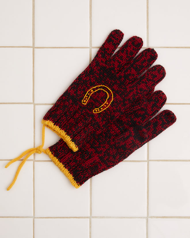 Set against a white tiled surface, the Melange Gloves are crafted from merino wool with a red and black speckled knit design, featuring yellow cuffs and an intricately embroidered horseshoe motif.