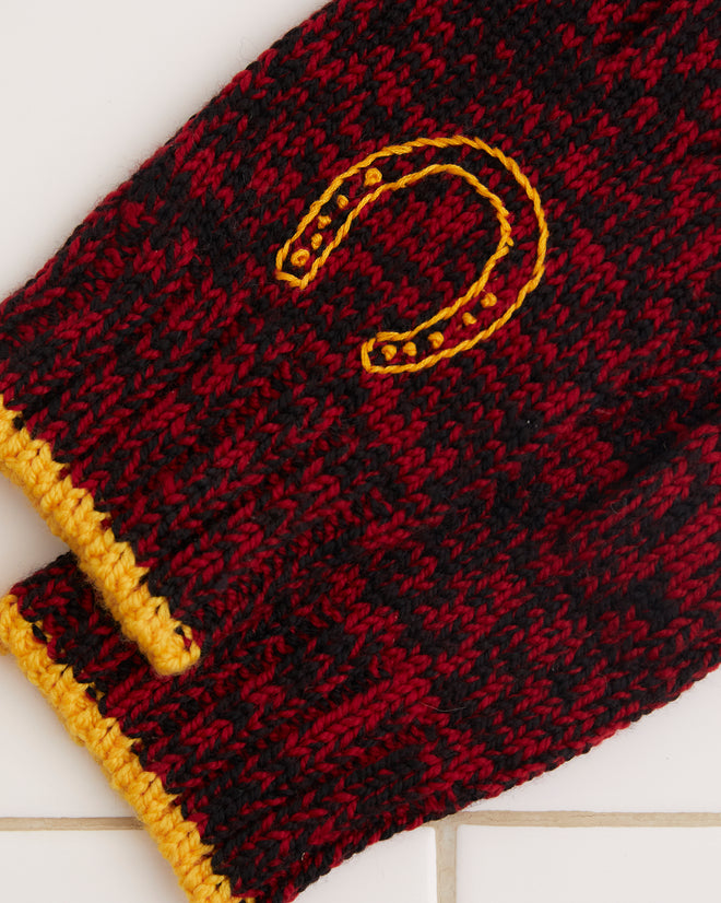 Set against a white tiled surface, the Melange Gloves are crafted from merino wool with a red and black speckled knit design, featuring yellow cuffs and an intricately embroidered horseshoe motif.