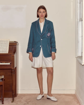 A woman stands indoors in the Fleur Souvenir Jacket, styled with white shorts and loafers, next to a piano with sheet music.