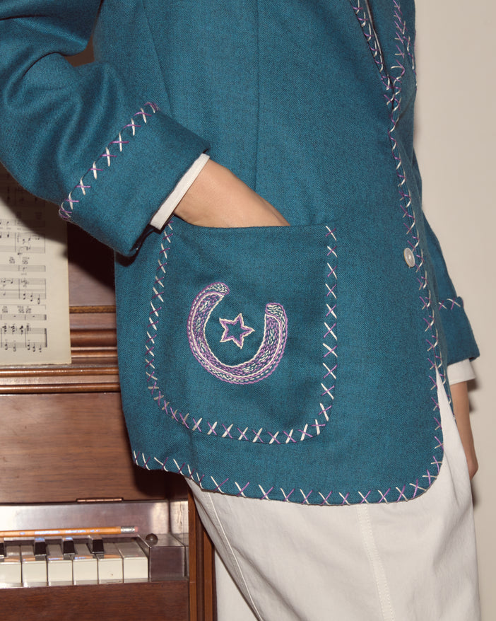 Detail of a women's jacket with a lavender horseshoe and star embroidered on a teal blue wool ground.