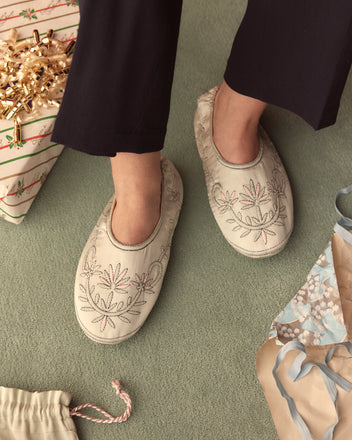 A person wearing Floral Souvenir Slippers stands on a green carpet, surrounded by wrapped gifts and curling ribbons.