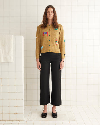 A woman stands in a tiled room wearing the Game Point Cardigan, a mustard-colored sweater adorned with hand-beaded patches, along with black high-waisted pants and black shoes. A towel hangs on the wall to her right.