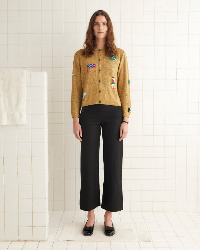 A woman stands in a tiled room wearing the Game Point Cardigan, a mustard-colored sweater adorned with hand-beaded patches, along with black high-waisted pants and black shoes. A towel hangs on the wall to her right.