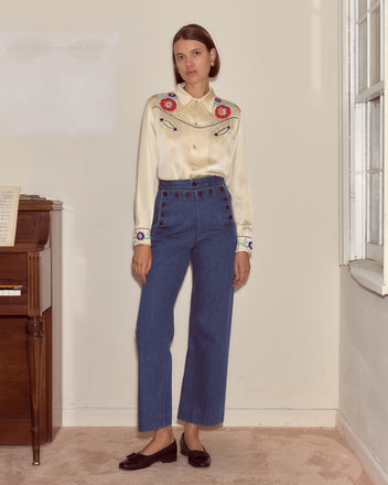 A woman wears the Garden Party Western Shirt with high-waisted blue jeans, standing indoors near a window and piano.