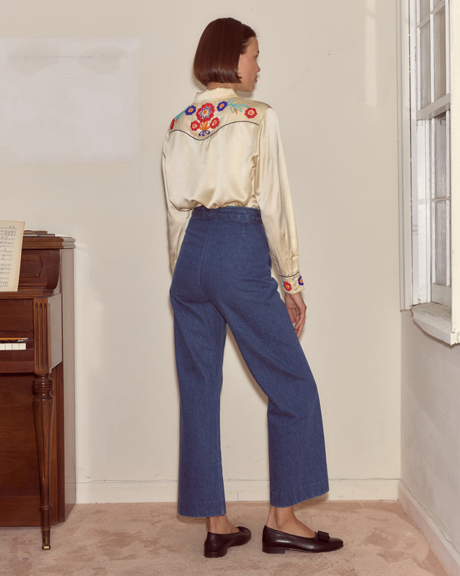 A woman wears the Garden Party Western Shirt, a cream beaded floral embroidered top, paired with blue jeans as she stands by a window and upright piano.