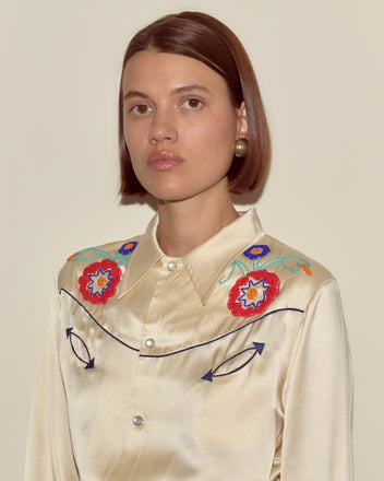 A woman wears the Garden Party Western Shirt, a cream silk top featuring colorful, beaded floral embroidery on the shoulders, set against a plain background.