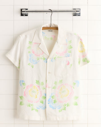 The Garden Stencil Shirt - S is a short-sleeve white button-up with a pastel floral print, evoking vintage damask style. Shown on a wooden hanger against a tiled wall.