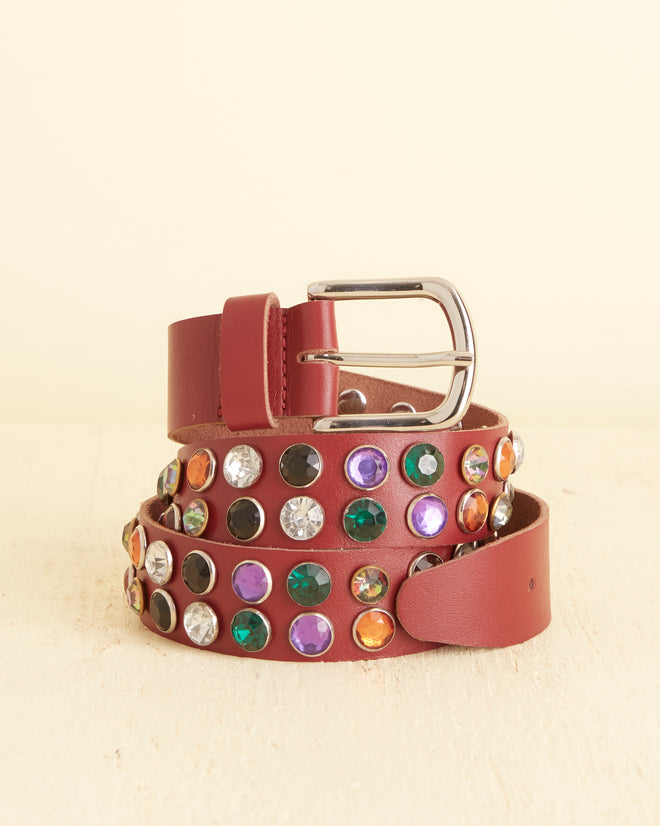 The Gem Stud Belt, a coiled red leather accessory adorned with various colored gemstones and featuring a metallic buckle, is displayed on a light-colored surface.