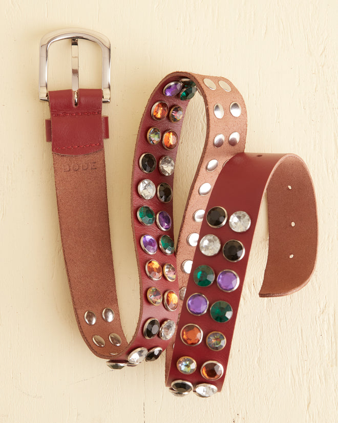 The Gem Stud Belt, a coiled red leather accessory adorned with various colored gemstones and featuring a metallic buckle, is displayed on a light-colored surface.