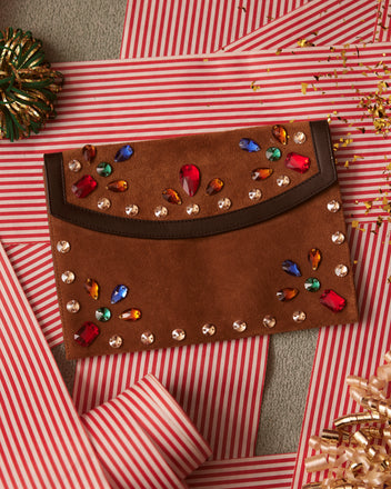 The Gem Venue Clutch in Brown/Black, an elegant suede envelope clutch embellished with jewels, rests on red and white striped paper, surrounded by festive ribbon and confetti.