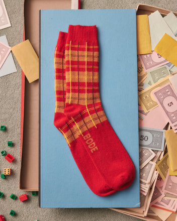 The County Plaid Socks - Red Multi, crafted from a luxurious blend of merino and cashmere, rest on a blue book, surrounded by Monopoly money and game pieces.