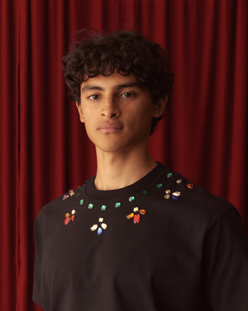 A person with curly hair, wearing the Gem Necklace Tee, stands against a red curtain.