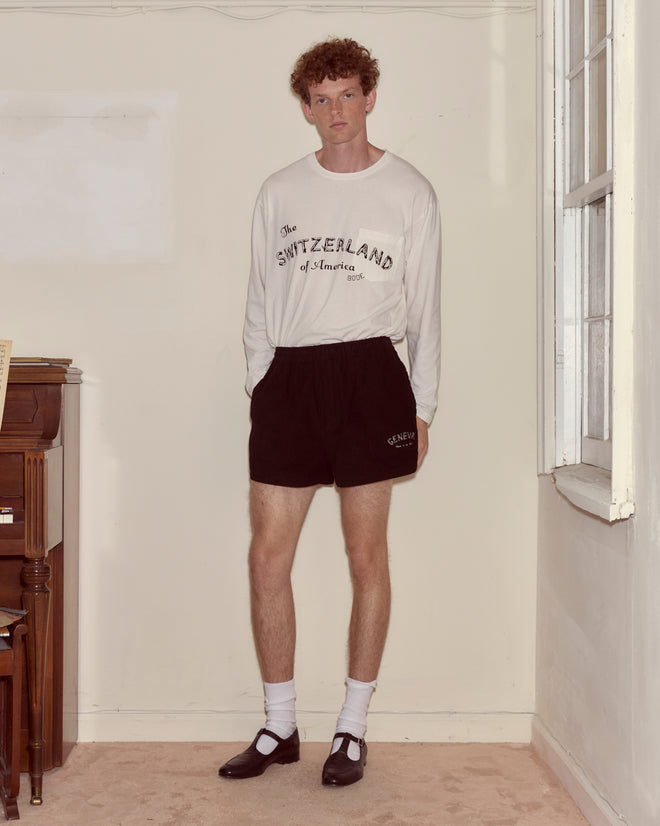 A man stands indoors in a white "Switzerland" shirt, Geneva Corduroy Shorts, white socks, and black Mary Jane shoes—a look reminiscent of Camp Geneva style.