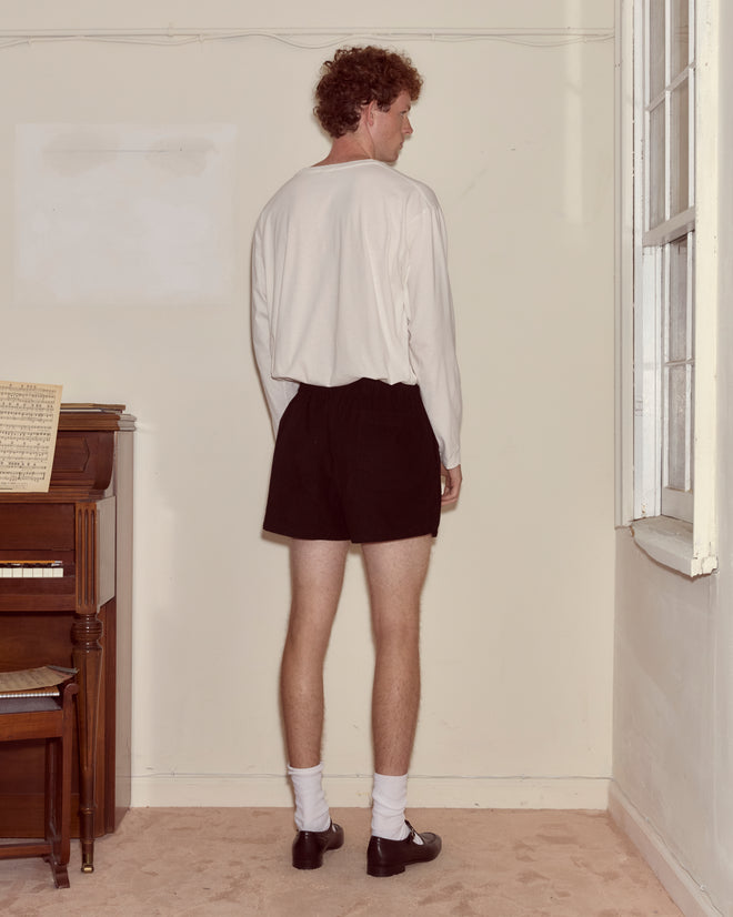 Someone in a white shirt and Geneva Corduroy Shorts, styled with a 1970s cut, stands facing a beige wall near a piano and window.