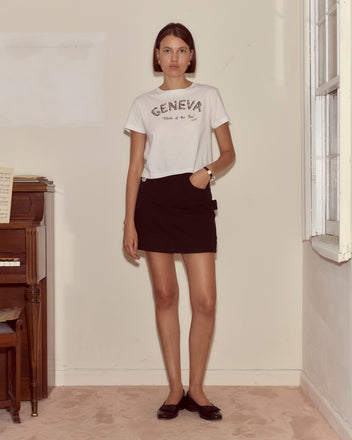 A woman in a fitted Geneva Tee and black skirt stands indoors beside a piano and window, gazing at the camera—evoking a scene from a vintage photo album.