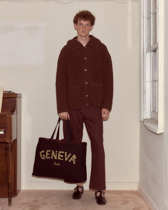 A person in a brown outfit holds a large black Geneva Tote, standing near a piano in a minimal room.