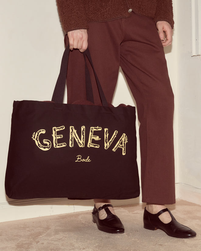 A person holds the Geneva Tote, a large black canvas bag inspired by Camp Geneva vintage photo albums, while wearing brown pants and black shoes.
