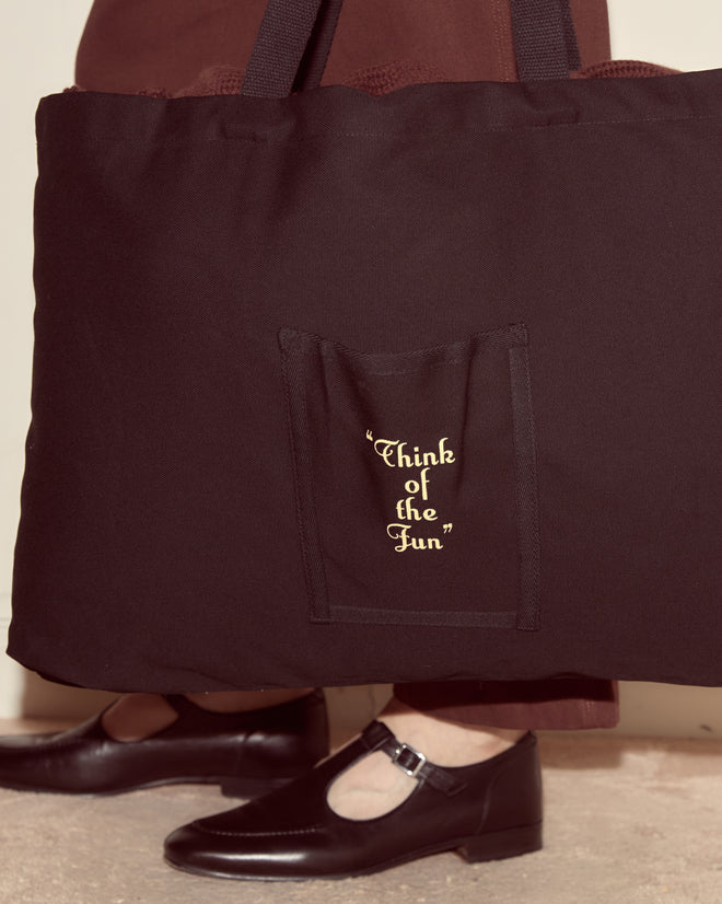 A person carrying the Geneva Tote, a black canvas bag with “Think of the Fun” in yellow on its pocket, inspired by a Camp Geneva vintage photo album.