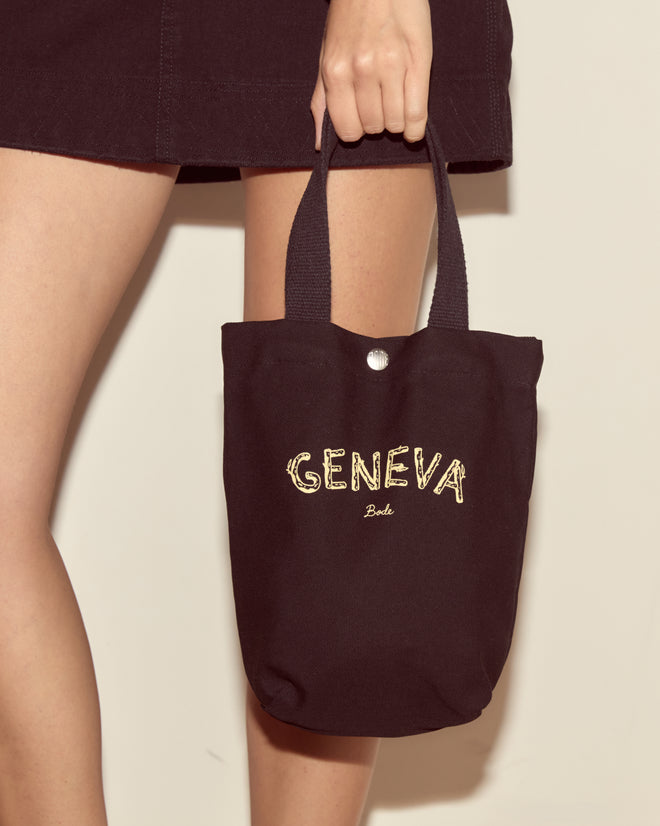 A person holds the Small Geneva Tote, featuring "GENEVA" in yellow text like a Camp Geneva keepsake, while wearing a dark skirt.