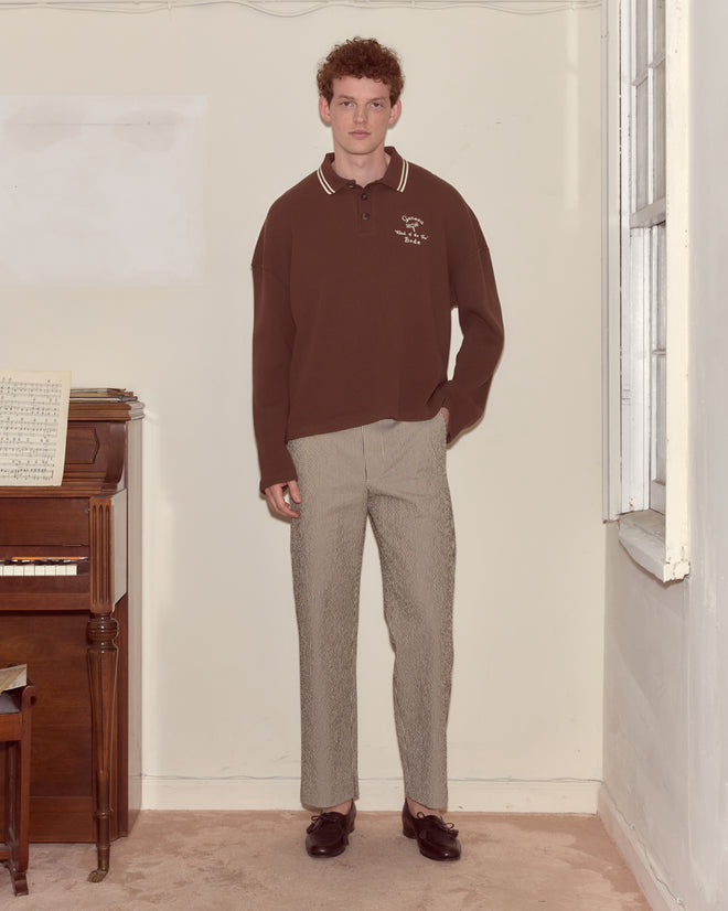 A person stands indoors by a piano, wearing a brown Geneva Waffle Polo and patterned pants.
