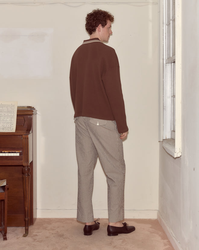 A man in a Geneva Waffle Polo and light pants stands with his back to the camera near a piano and window, capturing the cozy charm of Camp Geneva.