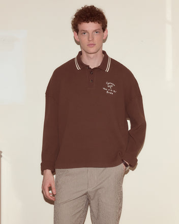 Young man with curly hair wears the Geneva Waffle Polo in brown and striped pants, standing against a plain light background.