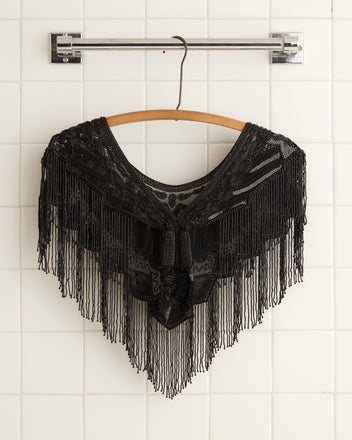 The Geode Collar - Black, an intricately patterned black shawl with subtle beadwork, is draped on a wooden hanger against a white tiled wall.