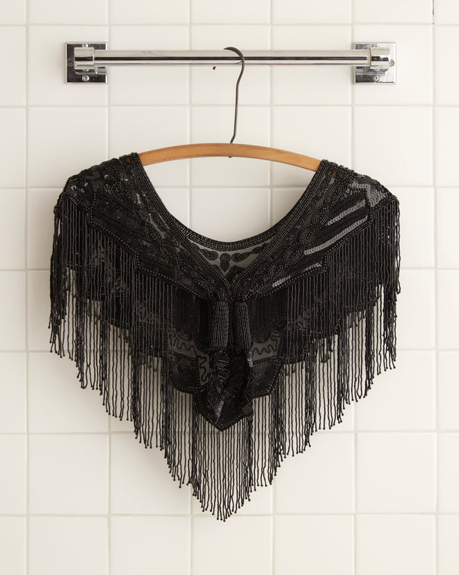 The Geode Collar - Black, an intricately patterned black shawl with subtle beadwork, is draped on a wooden hanger against a white tiled wall.