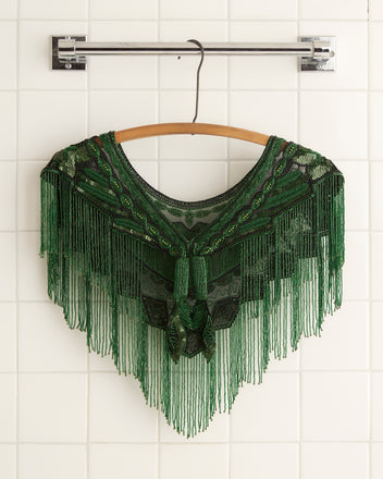 The Geode Collar - Green, with its intricate beadwork and fringe detail, hangs on a wooden hanger against a white tiled wall.