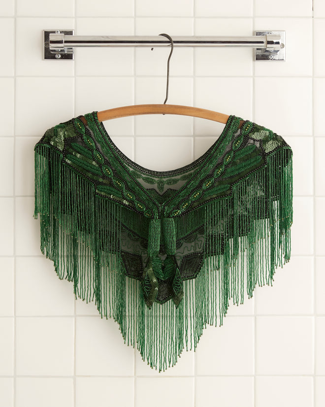 The Geode Collar - Green, with its intricate beadwork and fringe detail, hangs on a wooden hanger against a white tiled wall.